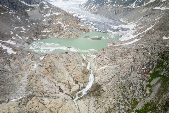 Aerial view of glacial meltwater cascading down rocky slopes into a serene lake, with the majestic Rh&ocirc;ne Glacier looming in the distance, Gletsch, Valais, Switzerland.