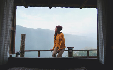 A woman sitting on wooden balcony with mountain views on foggy morning outside the window