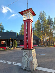 Tall red-and-white Arctic Circle marker stands on stone base beside a log cabin and forest. White painted line on pavement marks the latitude boundary under a partly cloudy northern sky. © Oskari