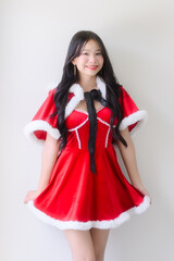 A young woman wearing a red santa dress with white fur trim and a black ribbon poses cheerfully against a plain white wall, smiling with festive charm in a bright indoor setting.