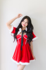 A young woman wearing a red santa dress with white fur trim and a black ribbon poses cheerfully against a plain white wall, smiling with festive charm in a bright indoor setting.
