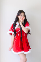 A young woman wearing a red santa dress with white fur trim and a black ribbon poses cheerfully against a plain white wall, smiling with festive charm in a bright indoor setting.