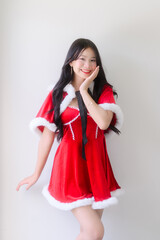 A young woman wearing a red santa dress with white fur trim and a black ribbon poses cheerfully against a plain white wall, smiling with festive charm in a bright indoor setting.