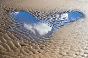 Heart-shaped small puddle on the North Sea beach of Norderney, reflecting white clouds in the blue sky. Tidal channel at low tide with typical sand structures that happen to form the symbol of love.