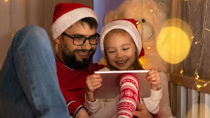 Happy family father and little daughter in red santa hats watch video on tablet for Christmas holidays. New Year Christmas. Computer games, surfing internet, social media