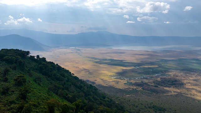 Aerial view of sunbeams piercing through clouds, illuminating the vast Ngorongoro Crater's diverse landscape with lush green slopes and golden plains, Ngorongoro Crater, Arusha Region, Tanzania.