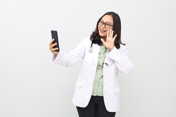 An Asian adult female doctor is standing in a doctor's coat, hanging a stethoscope around her neck while communicating online with a patient via mobile phone while waving her hand to greet.