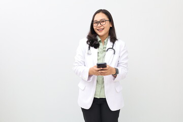 Asian adult female doctor standing while using mobile phone, wearing doctor's coat, hanging stethoscope around neck, looking at camera while smiling, sending message or using app.