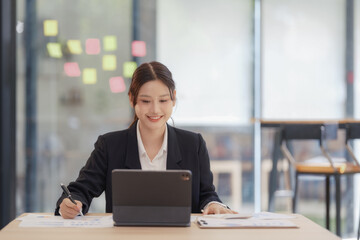 An Asian businesswoman smiling and working on a digital tablet at an office desk.