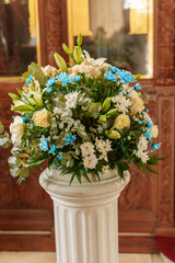 A vibrant floral arrangement featuring white lilies, blue and yellow flowers, and greenery stands in a classic white vase. This display is set against a richly designed wooden background.