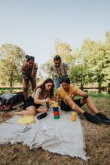 A cheerful group of friends plays a colorful block tower game on a blanket in a sunny park, with...