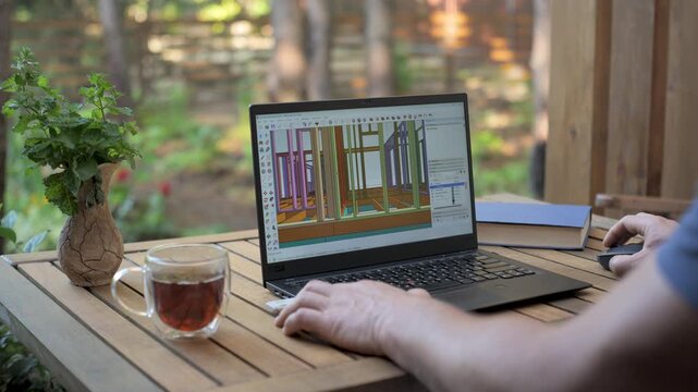 Architect working on a 3D model of an open-air house, a person using a laptop to design a 3D model of an open-air house, with tea, a book and a plant on a wooden table