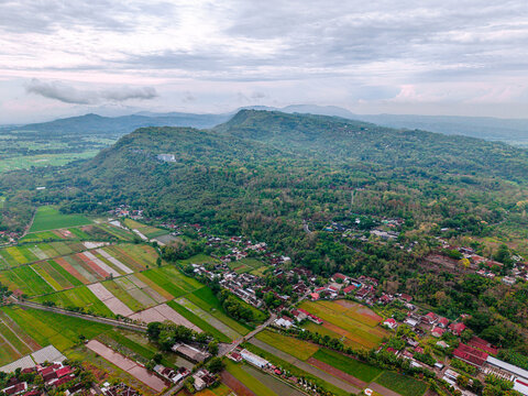 Aerial view of verdant rice fields meet the lush foothills under a vast, cloudy sky along Jalan Raya Solo - Yogyakarta, Yogyakarta, Daerah Istimewa Yogyakarta, Indonesia.
