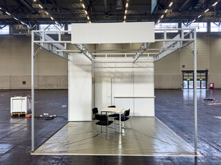 Weisser leerer Messestand in Messehalle mit schwarzen Stühlen und einem Tisch