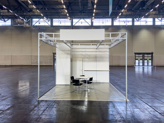 Weisser leerer Messestand in Messehalle mit schwarzen Stühlen und einem Tisch