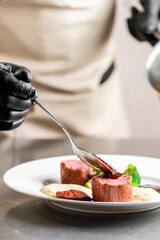 A chef in black gloves and a white apron plates a gourmet dish of seared meat with a puree and vegetables, drizzling sauce with a spoon