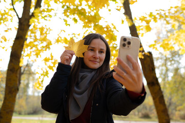 Smiling woman taking selfie with smartphone while holding yellow maple leaf against golden autumn trees background.