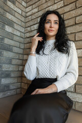Young woman sitting outdoors by a brick wall, dressed in a cozy white sweater and black skirt. Warm autumn sunlight creates a relaxed, stylish, and calm atmosphere.