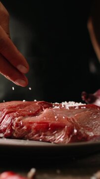 Vertical slow motion video chef hand seasoning raw beef steak with sea salt on kitchen table black background gourmet cooking concept fine dining premium meat restaurant preparation cinematic light