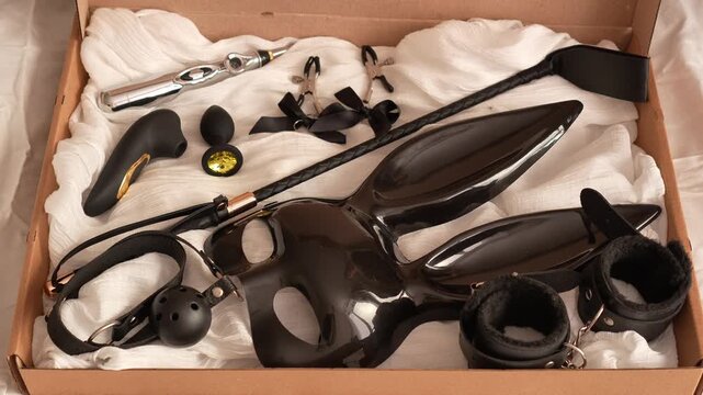 Woman in black transparent glove opens box with black adult toys. BDSM erotic set. Adult toys. Whip, gag, handcuffs, vacuum vibrator, butt plug, nipple clamps, electric stimulator, bunny mask