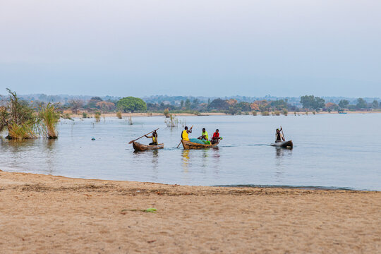 Ngala, Malawi - 28 September 2025: View of silhouetted figures rowing boats across the tranquil, glistening waters of Lake Malawi, under a soft, muted sky, from the sandy shore.