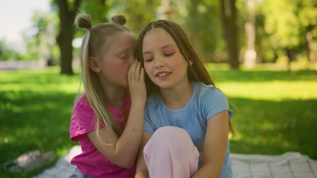 Cute little girl whispering secrets to her sister, having fun in green city park