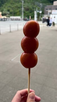 Vertical video of a female hand spins a stick with Mitarashi Dango (Japanese Rice Dumplings) outdoor at daytime with blurry background in Japan.