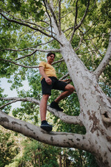 A man wearing a yellow shirt and black shorts climbs a wide tree in a green park. The moment...