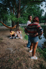 A group of friends spend a sunny day by a pond in a park. A hammock hangs between trees as others...