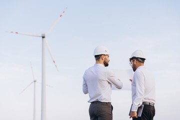 Engineers discussing blueprint in front of wind turbines, exploring sustainable energy solutions