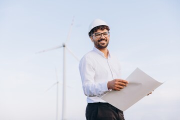 Arabian engineer holding blueprint inspecting wind turbines in field