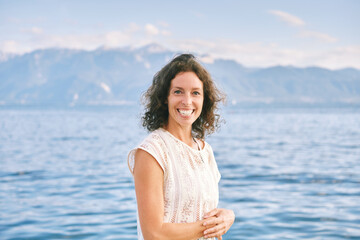 Outdoor close up portrait of happy candid woman, brown curly hair, big smile, posing with lakeside nature background