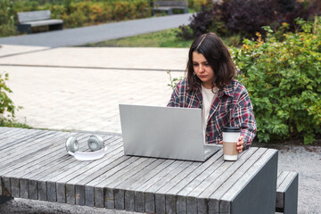 Young woman sitting outdoors at a wooden table with laptop coffee cup, wearing a plaid shirt and focusing on her work. Headphones lying beside her. Concept of remote work, freelancing, studying online