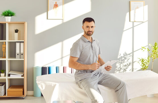 Young confident doctor physiotherapist sitting on couch in rehab clinic. Portrait of male professional physical therapist looking at camera in rehabilitation hospital . Physiotherapy concept - Powered by Adobe