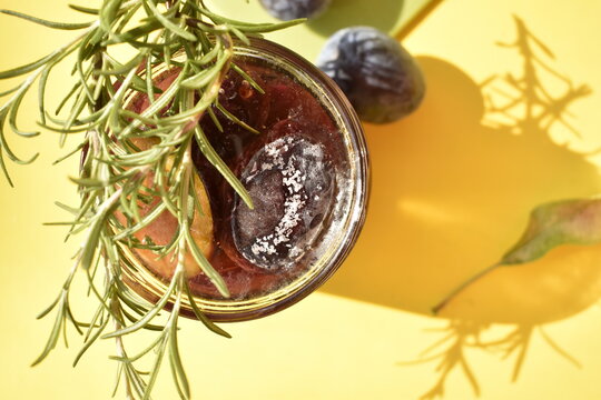 open jar of preserved plums in sugar decorated by rosemary sprig and whole plums on yellow background, top view