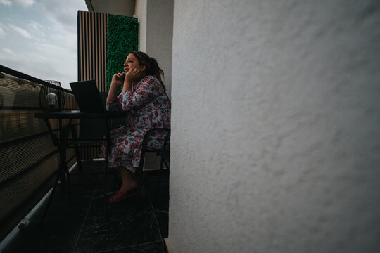 A woman sits at a balcony desk with a laptop, talking on a phone, wearing a floral dress. She appears focused and relaxed, blending work with personal life in a casual outdoor setting.