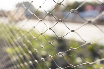 Close-up of a metallic wire fence with a blurred background of greenery and soft sunlight, creating a serene atmosphere and highlighting the texture of the fence
