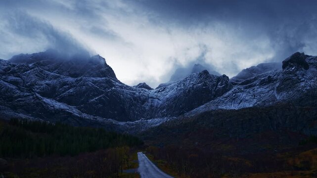 Cars on narrow country road through the forest by snow covered mountains on a cloudy dark autumn evening. Time Lapse. Lofoten Islands, Northern Norway.