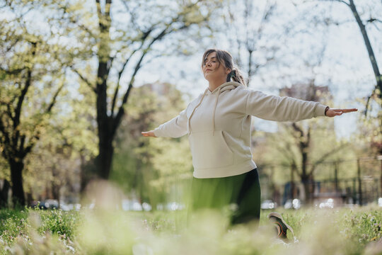 A woman wearing a hoodie stretches her arms outward while kneeling on a grassy area under sunlight in a peaceful park. - Powered by Adobe