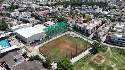 Aerial drone shot of a sports complex featuring a cricket pitch and netted practice areas, surrounded by dense urban residential buildings on a sunny day.