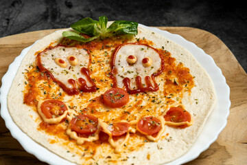 Close-up of Halloween-themed pizza with skull-shaped eyes and tomato mouth on white plate over wooden board, top view food photography for spooky presentation