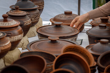 A woman&rsquo;s hand gently touches a lidded handmade ceramic jar at a rustic pottery market, surrounded by earthy-toned, intricately carved artisanal cookware and bowls.