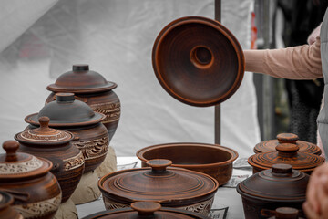 A woman&rsquo;s hand lifts a lid from a handmade ceramic pot at a rustic pottery market, surrounded by earthy-toned, intricately carved artisanal jars and bowls.