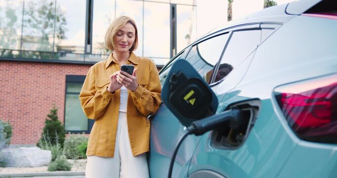 Woman using smartphone app while standing next to her electric car charging at a portable charging station on a city street.