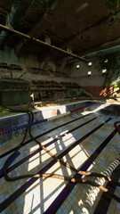 Sunlight streams into a deserted indoor pool, highlighting the faded tiles and remnants of seating. Plants creep through cracks, reclaiming a space once full of life and laughter.