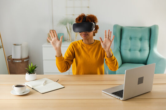 African American young woman wearing using virtual reality metaverse VR glasses headset at home. Girl touching air during VR experience on virtual reality helmet. Simulation hi-tech videogame concept