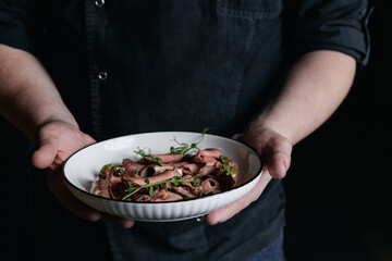 Male chef hands holding roast beef salad in white bowl on dark background