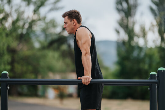 A man in a black sleeveless tank exercises on outdoor parallel bars, focusing on upper body strength. The park setting features greenery and a calm, sunny mood.