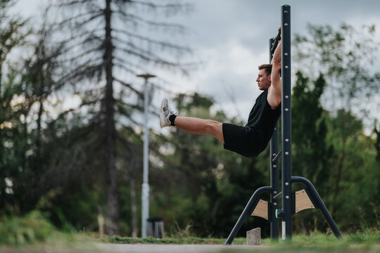 A fit man in black athletic wear uses a vertical leg raise station in a park, demonstrating outdoor strength training. Overcast sky and trees frame a calm fitness moment.