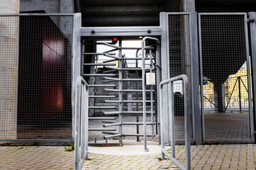 A metal turnstile gate stands at the entrance of a modern building. The surrounding area features concrete walls and a paved pathway.